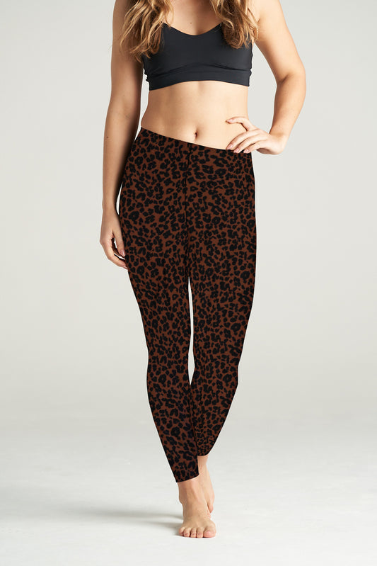 Woman standing barefoot in a relaxed pose wearing high-waisted brown cheetah print leggings and a black sports bra, modeling the leggings in a studio setting.