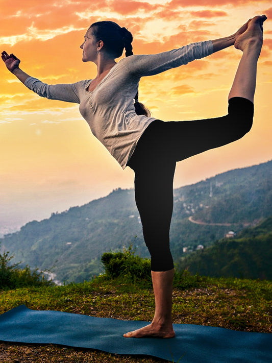 Woman practicing yoga outdoors at sunrise on a grassy hilltop, wearing black capri leggings and a long-sleeve top, standing in dancer's pose on a blue yoga mat with mountains in the background.