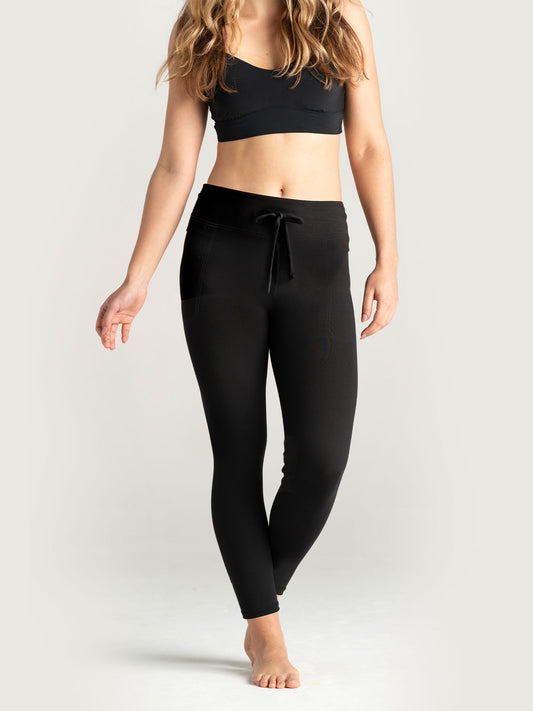 Woman wearing black lounge leggings with a drawstring waistband and side pockets, paired with a black sports bra, posing barefoot in a neutral studio setting