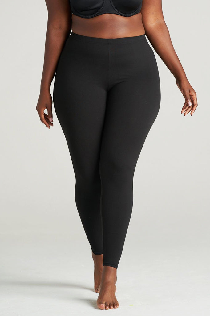 Plus-size woman modeling full-length black leggings in a studio setting, barefoot, with a confident walking pose and wearing a black bra