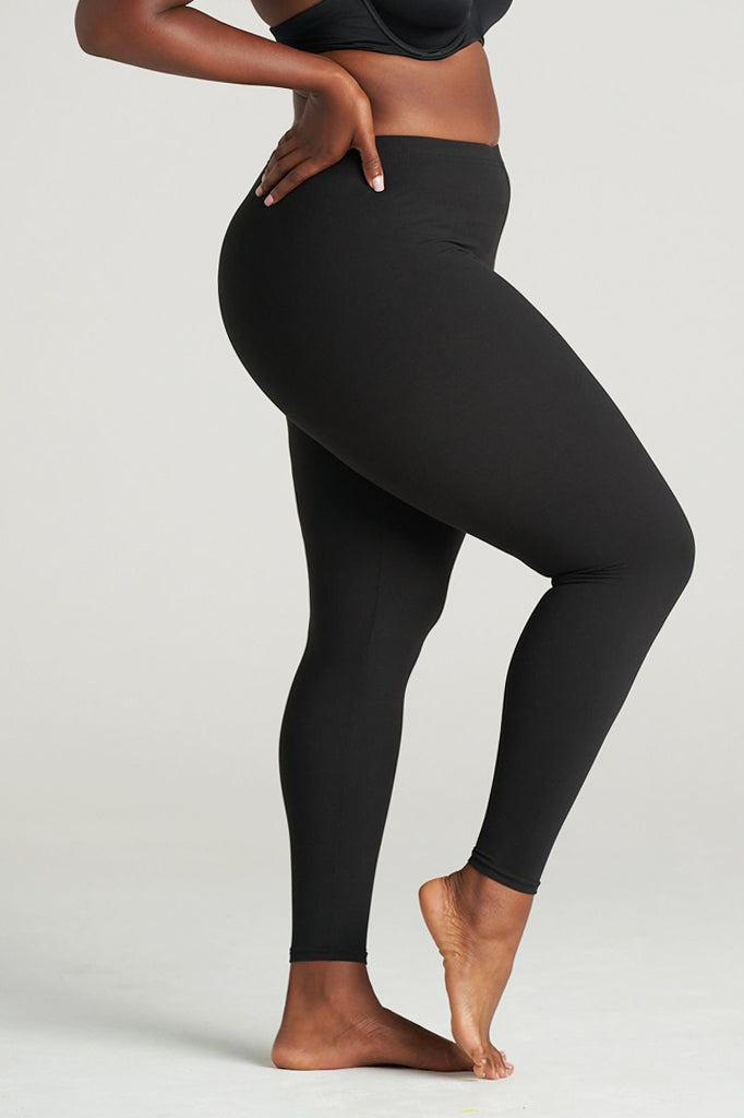 Side profile of a plus-size woman modeling full-length black leggings, barefoot, with one leg slightly lifted and one hand on her waist, showcasing the comfortable stretch and curve-hugging fit