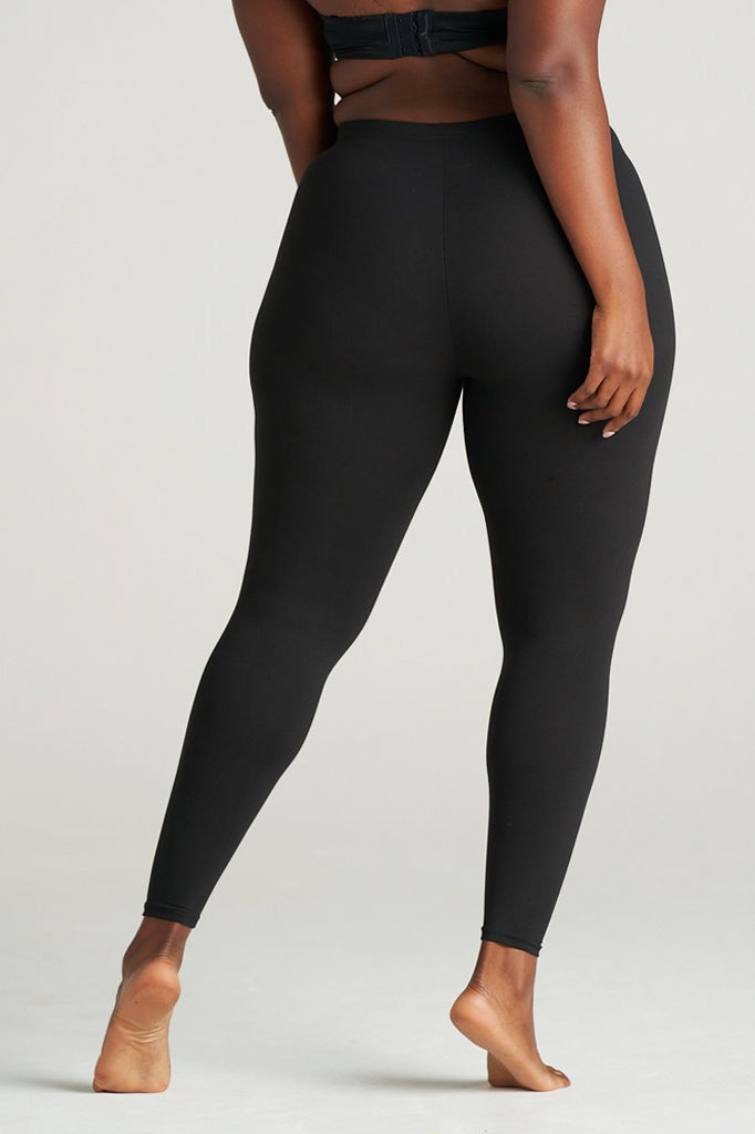 Rear view of a plus-size woman modeling full-length black leggings, barefoot, with one leg slightly lifted, highlighting the smooth fit and contouring fabric around the hips and legs.