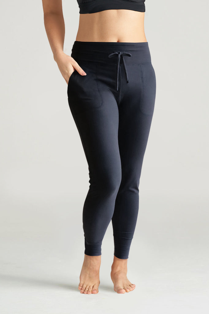 Woman wearing black lounge leggings with a drawstring waistband and side pockets, paired with a black sports bra, posing barefoot in a neutral studio setting.