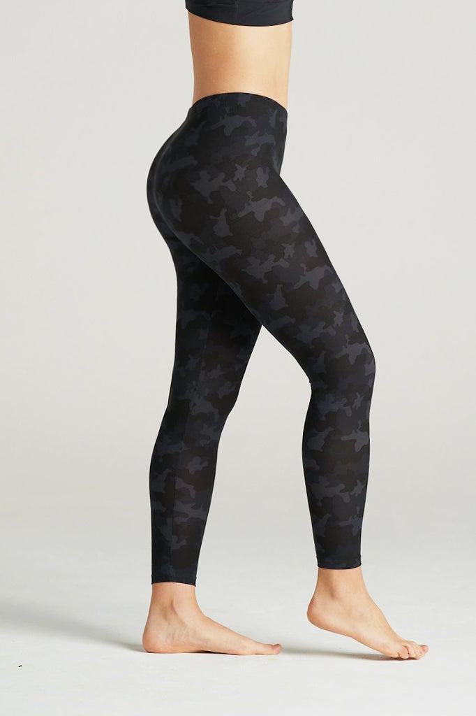 Woman wearing high-waisted black camouflage leggings and a black sports bra, posing barefoot in a minimal indoor studio. These muted black camo print leggings feature a sleek, body-contouring fit perfect for yoga, gym workouts, or athleisure styling. Designed for comfort, flexibility, and a flattering silhouette.