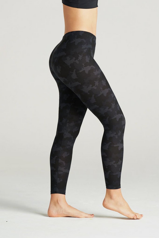 Woman wearing high-waisted black camouflage leggings and a black sports bra, posing barefoot in a minimal indoor studio. These muted black camo print leggings feature a sleek, body-contouring fit perfect for yoga, gym workouts, or athleisure styling. Designed for comfort, flexibility, and a flattering silhouette.