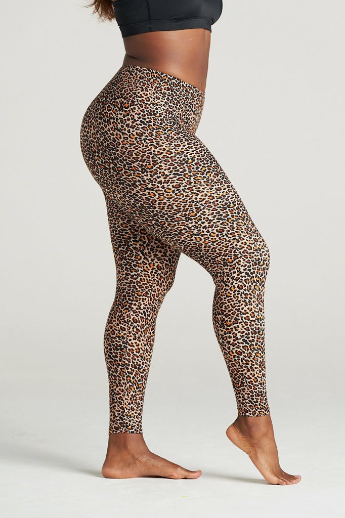 Plus-size woman wearing high-waisted brown leopard print leggings paired with a black sports bra. Comfortable and stretchy animal print leggings perfect for workouts, yoga, or casual wear. Full-length design with a body-hugging fit, photographed barefoot in a studio with a neutral background.