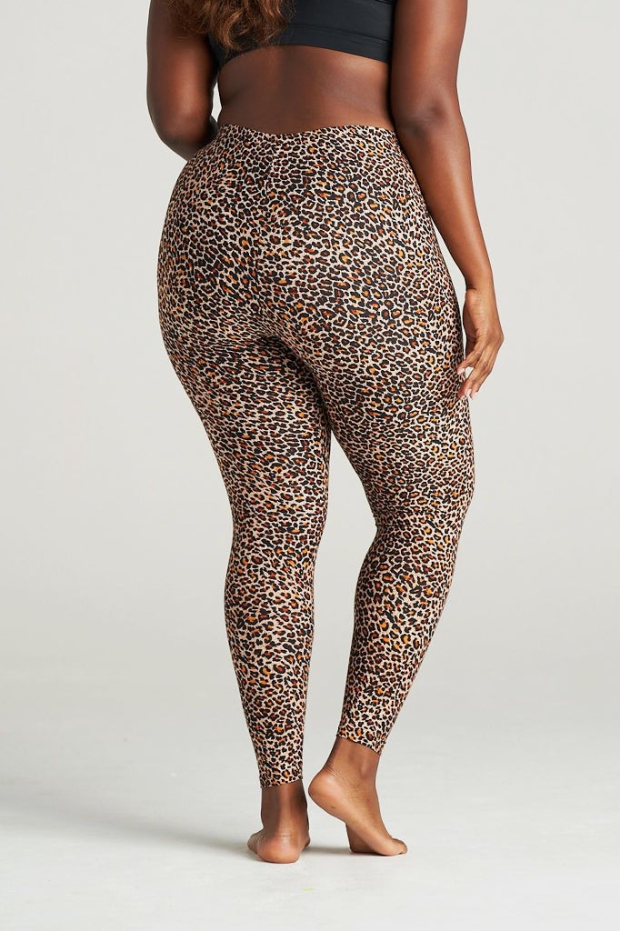 Plus-size woman wearing high-waisted brown leopard print leggings paired with a black sports bra. Comfortable and stretchy animal print leggings perfect for workouts, yoga, or casual wear. Full-length design with a body-hugging fit, photographed barefoot in a studio with a neutral background.