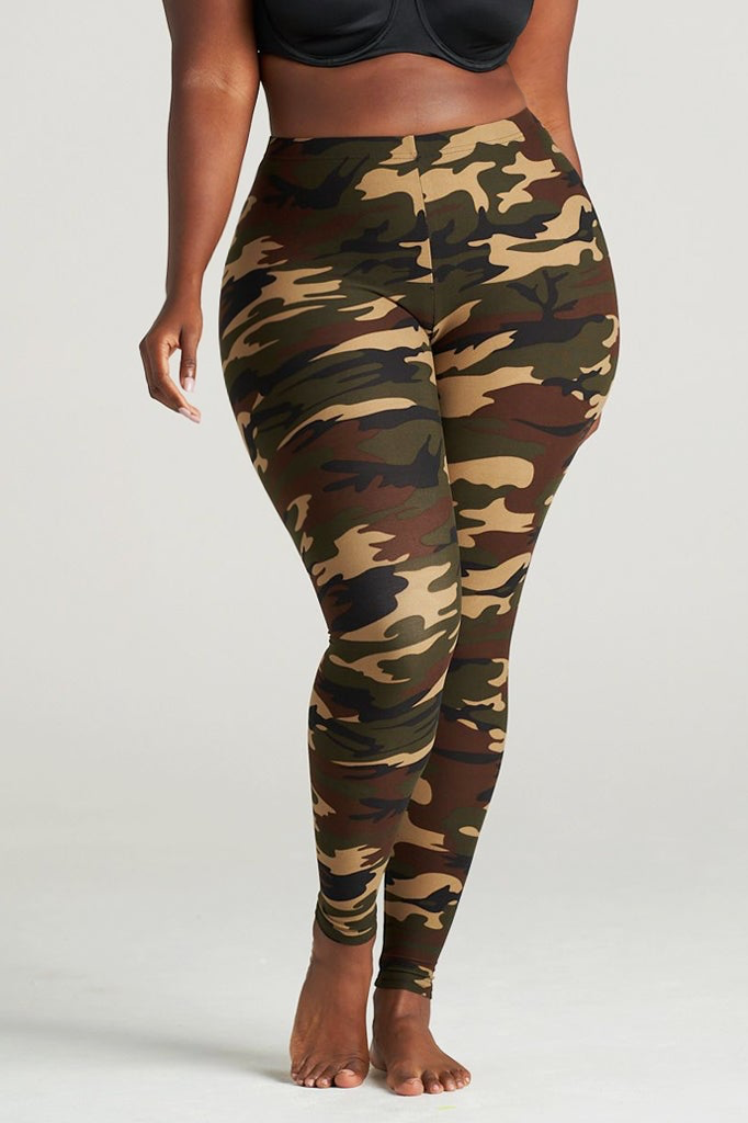 Plus Size Woman modeling high-waisted green camouflage leggings, barefoot in a neutral indoor studio. The leggings feature an earthy camo pattern in shades of olive, brown, and khaki, paired with a black sports bra for a sporty and confident look. Ideal for gym, yoga, or casual streetwear.