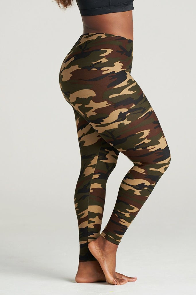 Plus Size Woman modeling high-waisted green camouflage leggings, barefoot in a neutral indoor studio. The leggings feature an earthy camo pattern in shades of olive, brown, and khaki, paired with a black sports bra for a sporty and confident look. Ideal for gym, yoga, or casual streetwear.