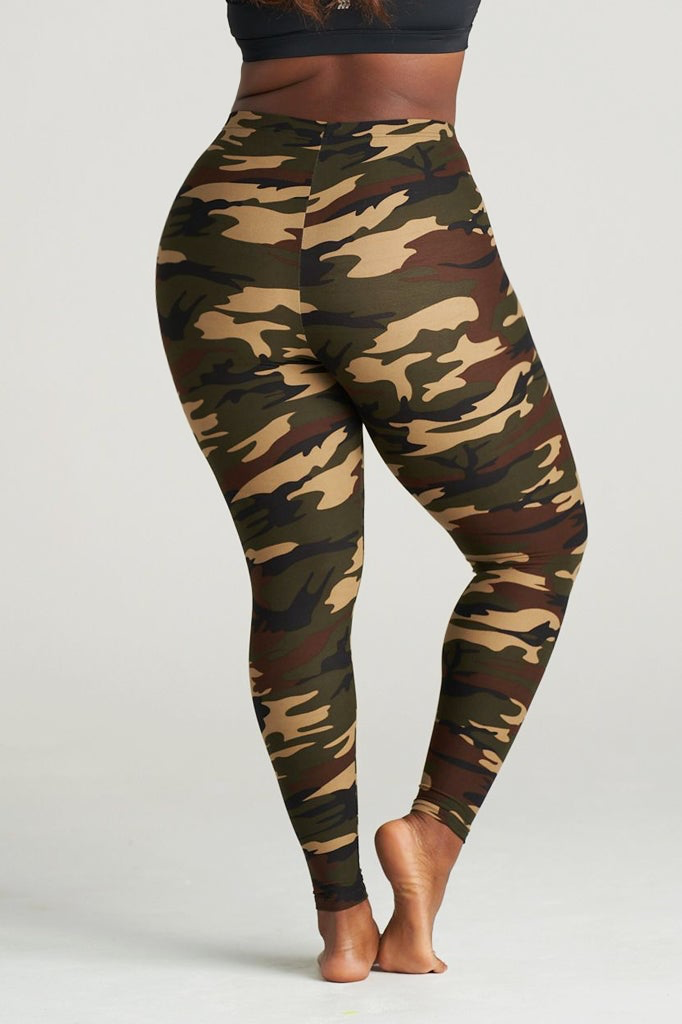 Plus Size Woman modeling high-waisted green camouflage leggings, barefoot in a neutral indoor studio. The leggings feature an earthy camo pattern in shades of olive, brown, and khaki, paired with a black sports bra for a sporty and confident look. Ideal for gym, yoga, or casual streetwear.