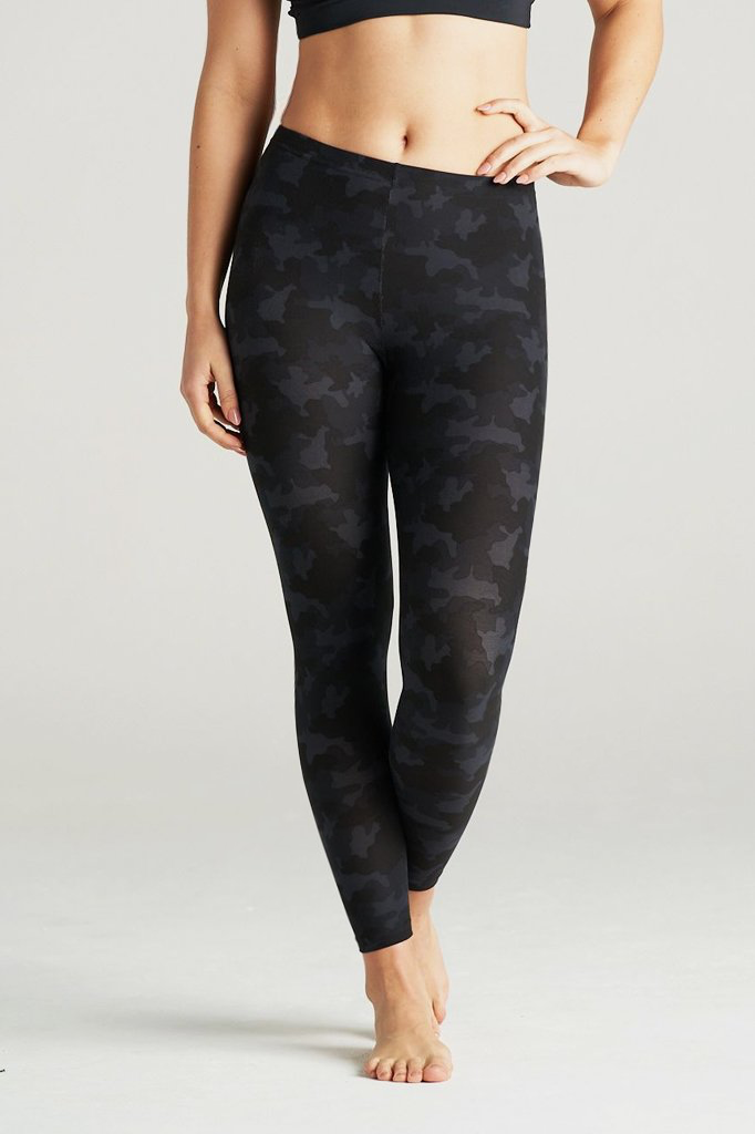 Woman wearing high-waisted black camouflage leggings and a black sports bra, posing barefoot in a minimal indoor studio. These muted black camo print leggings feature a sleek, body-contouring fit perfect for yoga, gym workouts, or athleisure styling. Designed for comfort, flexibility, and a flattering silhouette.