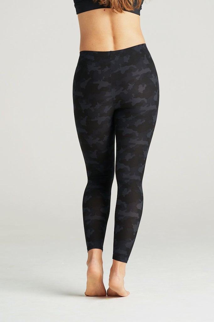 Woman wearing high-waisted black camouflage leggings and a black sports bra, posing barefoot in a minimal indoor studio. These muted black camo print leggings feature a sleek, body-contouring fit perfect for yoga, gym workouts, or athleisure styling. Designed for comfort, flexibility, and a flattering silhouette.