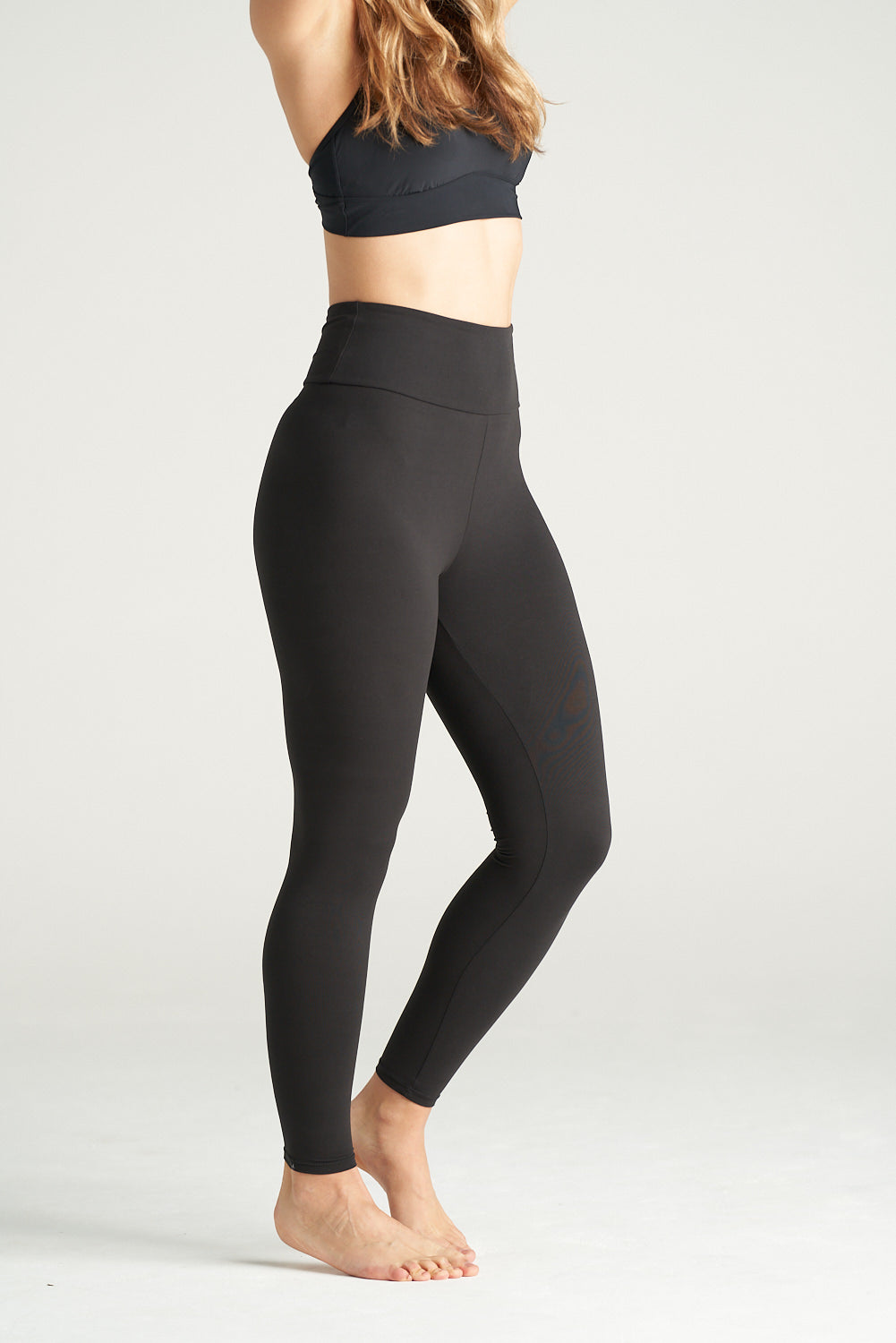 Woman standing in a relaxed side profile pose wearing black high-waisted full-length leggings and a black sports bra, showcasing activewear against a neutral background.