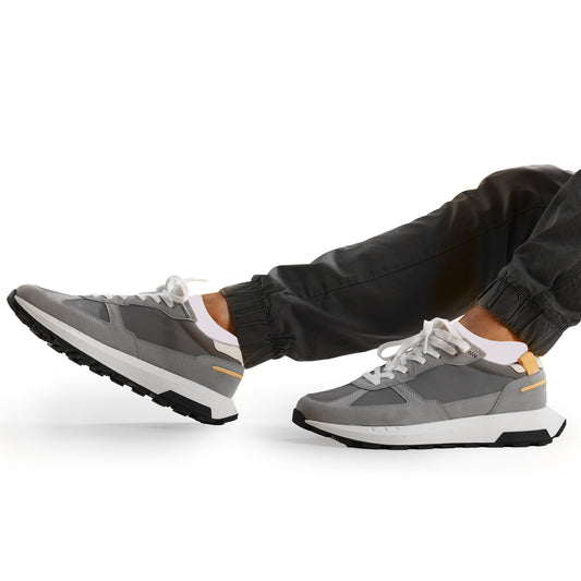 Man seated in a relaxed pose wearing white cushioned low-cut socks and modern gray athletic sneakers with white soles and yellow accents, paired with charcoal jogger pants